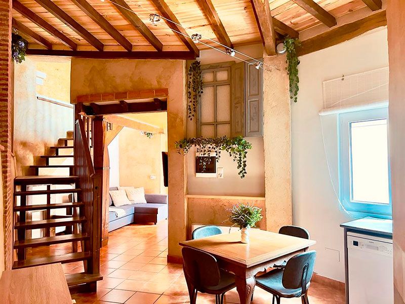 Comedor con mesa cuadrada, escaleras y salón con sofá al fondo